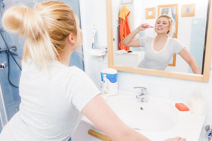 blond woman with messy bun of hair, looking in mirror brushing teeth