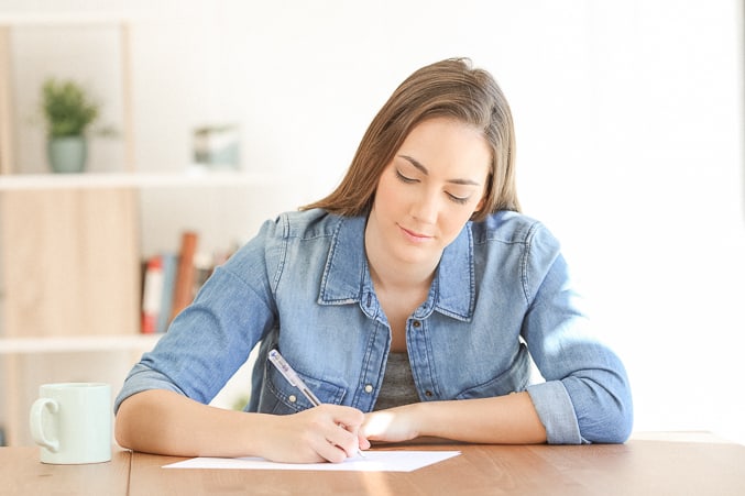 woman wearing a blue shirt, writing on a piece of paper on a brown wood table