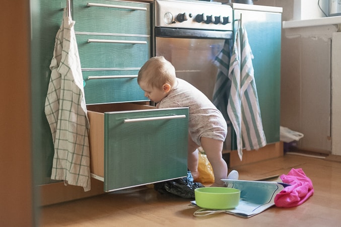 toddler boy reaching into open, green coloured drawer in kitchen. Pile of dishes on floor beside him