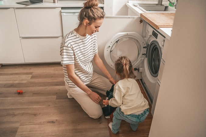 Mom kneeling on the floor beside washing machine, teaching child how to load washer.